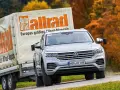 Srebrny Volkswagen Touareg holuje przyczepę z logo "Auto Bild allrad", jadąc po krętej drodze wśród jesiennych drzew.