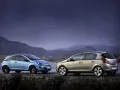 Dwa samochody Opel Corsa D, jeden niebieski, drugi beżowy, stoją na kamiennym murze nad wodą.