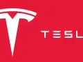 Biały, stylizowany symbol litery "T" i napis "TESLA" na czerwonym tle. Tak wygląda znaczek Tesli.