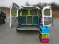 Tani bus do 10 tys. z otwartym tyłem, w którym widać zielone i niebieskie skrzynki. Obok stos kolorowych skrzynek.