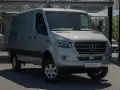 Srebrny bus Mercedes-Benz Sprinter, idealny jako jaki bus na budowę, stoi na parkingu przed nowoczesnym budynkiem.