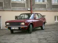 Czerwony fiat 128, klasyk z lat 70., zaparkowany na brukowanym dziedzińcu przed budynkiem.