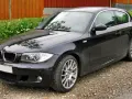 Czarny BMW e81, lśniący po deszczu, stoi na żwirowej drodze obok zielonego żywopłotu i drewnianego ogrodzenia.