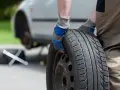 Mężczyzna w rękawiczkach trzyma koło zapasowe, przygotowując się do jego zamocowania w bagażniku. Obok leży klucz.