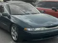 Subaru SVX w ciemnozielonym kolorze, z charakterystycznymi oknami w stylu "glass-canopy", zaparkowany obok czerwonego samochodu.
