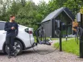 Mężczyzna z telefonem przy białym samochodzie elektrycznym, który ładuje się obok stojaka z rowerami. Obok stoją dwie ładowarki do samochodów elektrycznych.