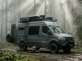 Szary bus z napędem 4x4, gotowy na przygodę w lesie. Na dachu bagażnik z wyposażeniem, z boku drabinka i koło zapasowe.