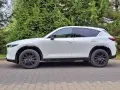 Nowa Mazda CX-5 w białym kolorze, zaparkowana na brukowanym podjeździe, otoczona zielenią drzew.