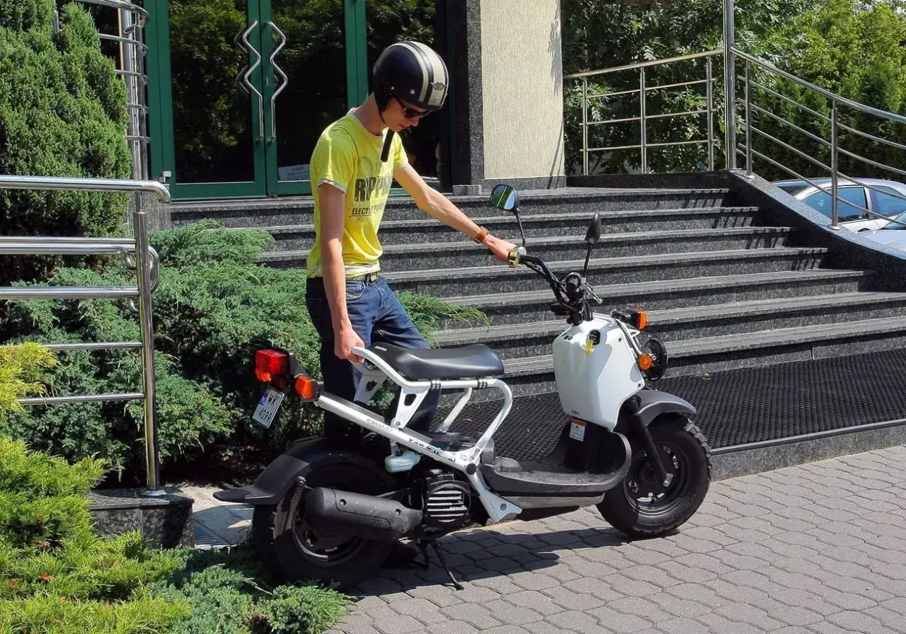 Chłopak w kasku i żółtej koszulce przy białym skuterze. Zastanawiasz się, ile kosztuje karta motorowerowa?
