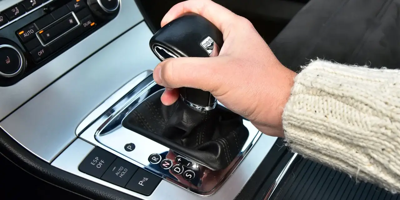 Dłoń trzyma gałkę zmiany biegów w samochodzie. Widoczna jest automatyczna skrzynia biegów DSG z przyciskami ESP, Auto Hold i czujnikami parkowania.