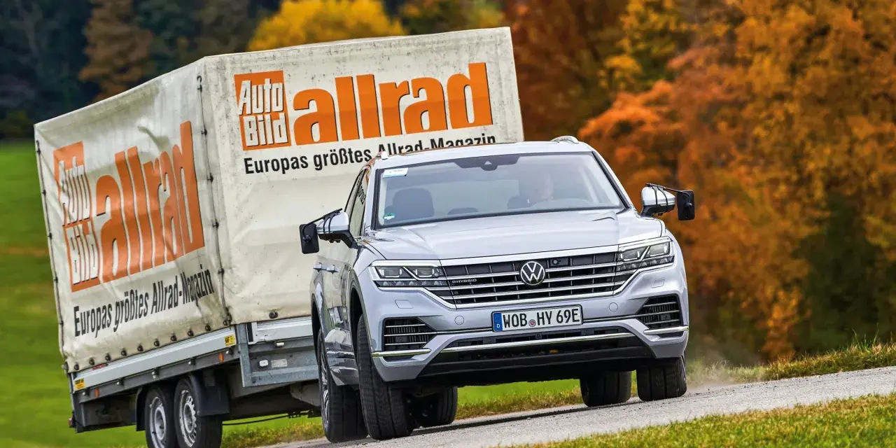 Srebrny Volkswagen Touareg holuje przyczepę z logo "Auto Bild allrad", jadąc po krętej drodze wśród jesiennych drzew.