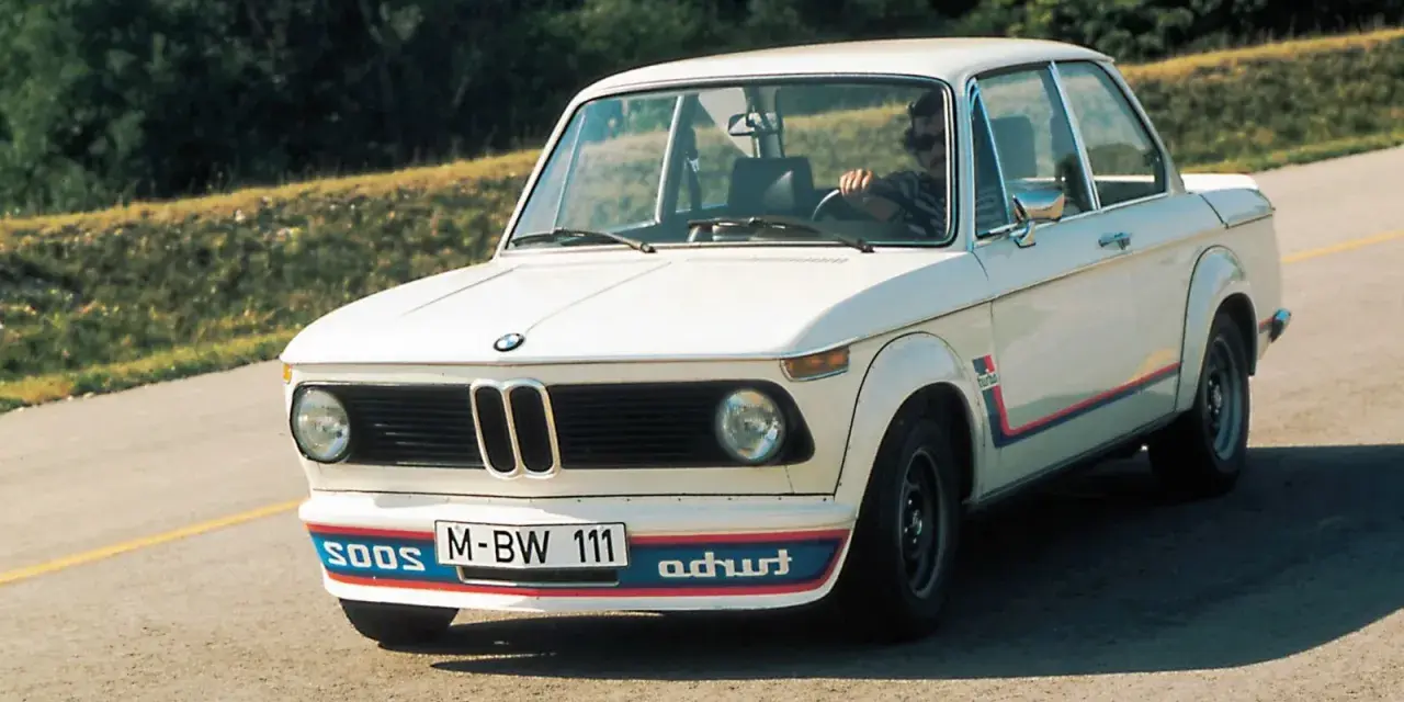 Białe BMW E20 z kolorowymi pasami i napisem "turbo" na błotniku, z kierowcą za kierownicą, jedzie po asfaltowej drodze.