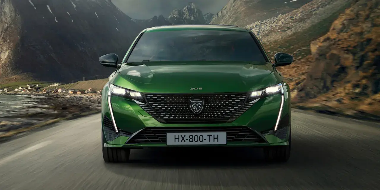 Zielony Peugeot 308 T7 jedzie drogą w malowniczym, górskim krajobrazie z widokiem na małą wioskę nad wodą.