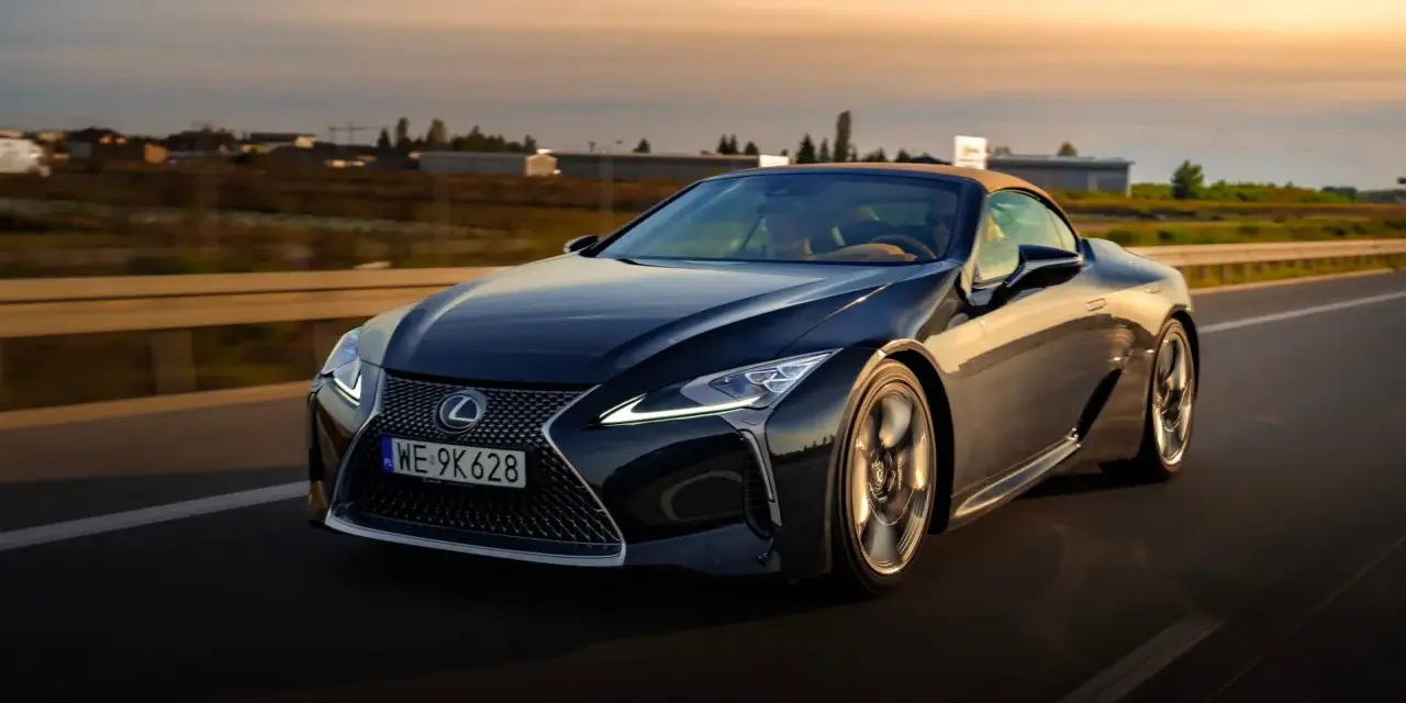 Ciemny Lexus LC 500 z beżowym dachem sunie po drodze podczas zachodu słońca. Sprawdź jego lexus lc 500 cena.