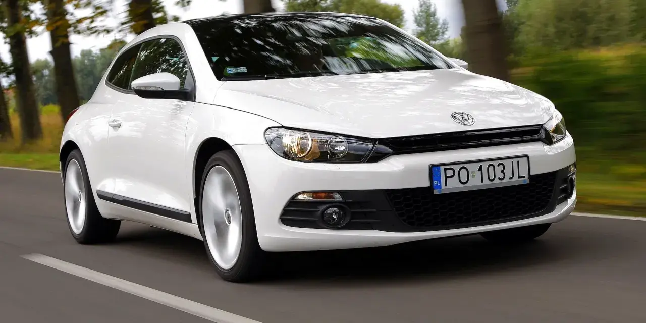 Biały Volkswagen Scirocco pędzi po drodze wśród drzew.