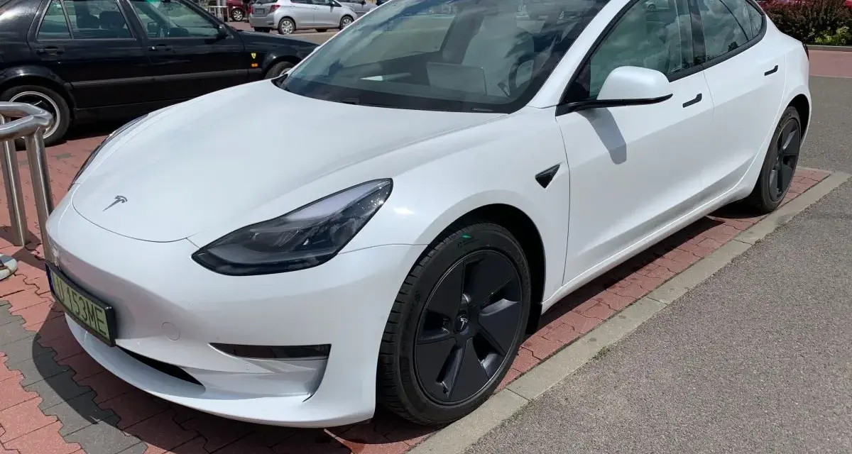 Biała Tesla Model 3 na parkingu przed sklepem. Zastanawiasz się, ile kosztuje najdroższa Tesla? Ten model to marzenie wielu kierowców.