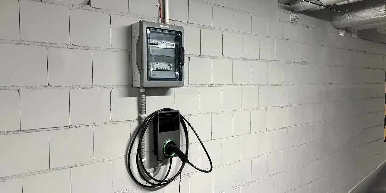 Nowoczesne gniazdo do ładowania samochodu elektrycznego Autel zamontowane na białej, ceglanej ścianie obok skrzynki z bezpiecznikami.