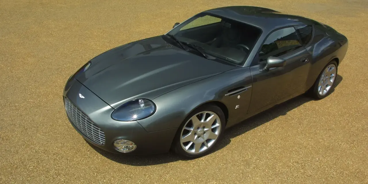 Szary Aston Martin DB9 na żwirowym podjeździe. Zastanawiasz się, ile kosztuje Aston Martin? To auto to marzenie wielu.