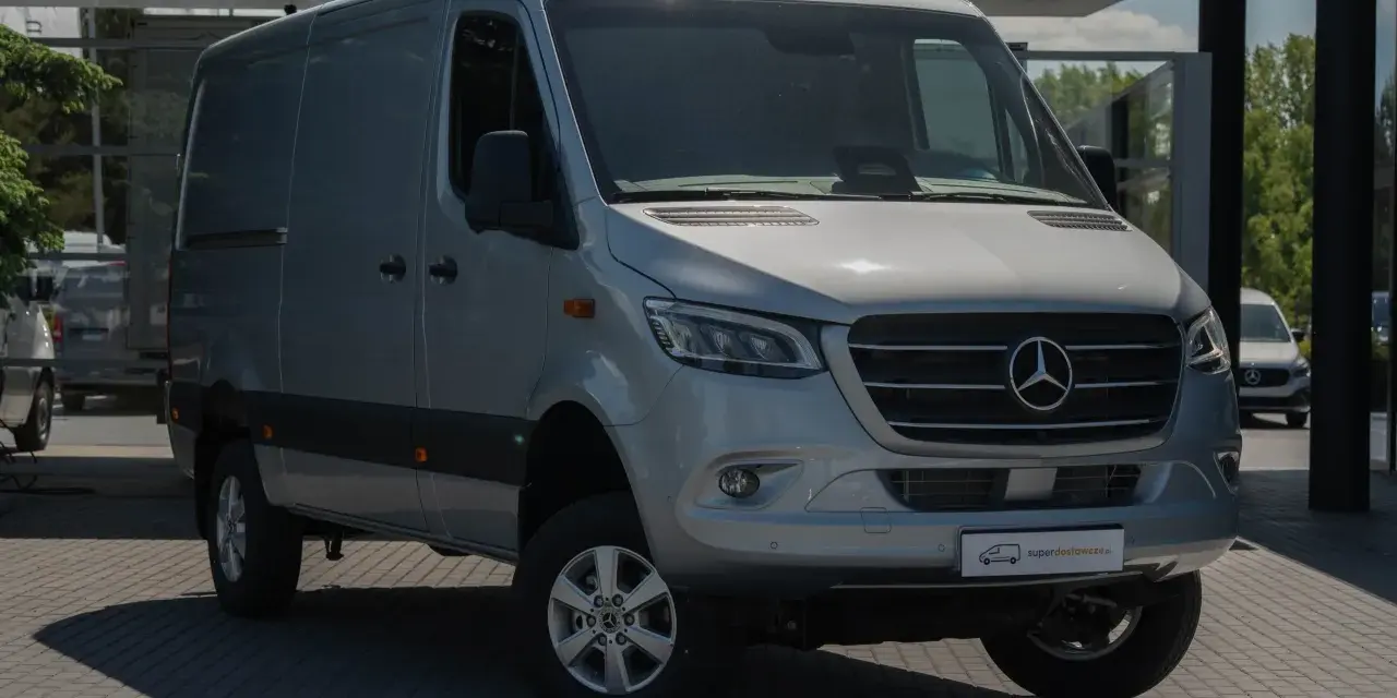 Srebrny bus Mercedes-Benz Sprinter, idealny jako jaki bus na budowę, stoi na parkingu przed nowoczesnym budynkiem.