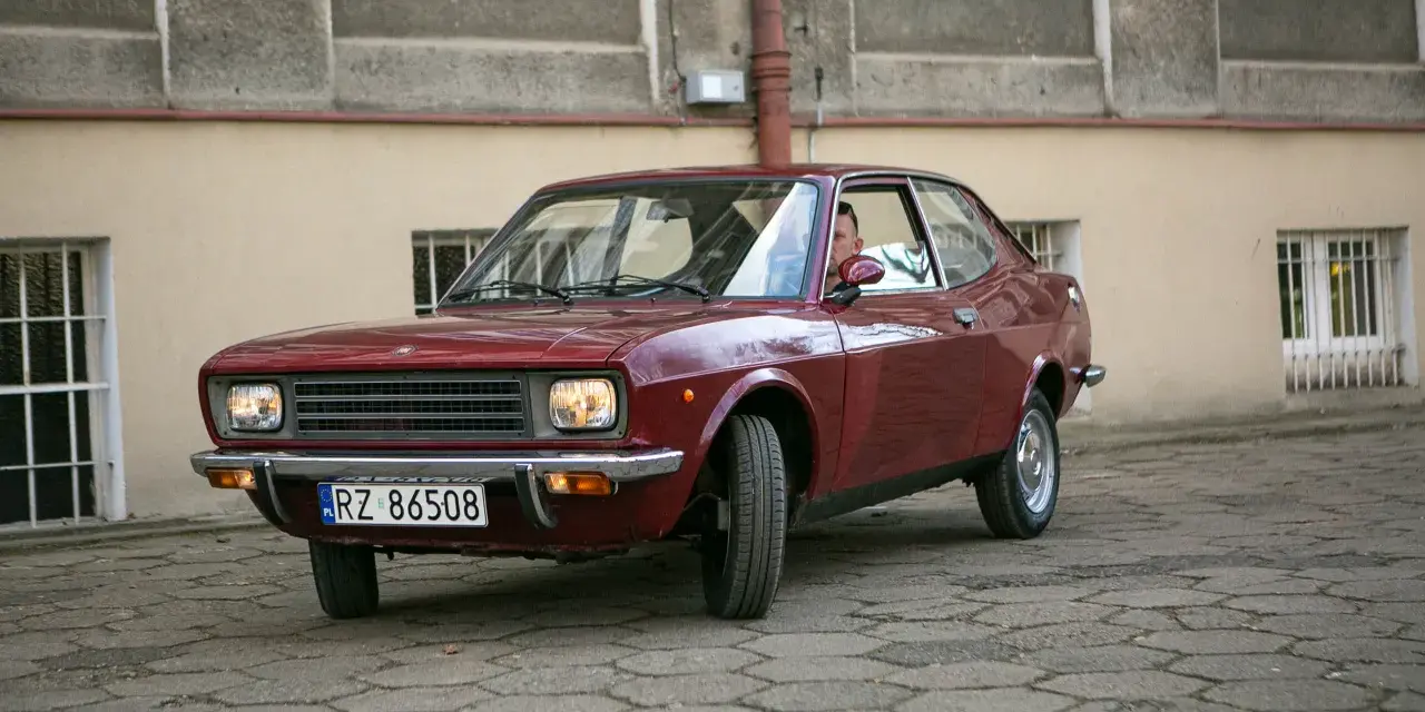 Czerwony fiat 128, klasyk z lat 70., zaparkowany na brukowanym dziedzińcu przed budynkiem.