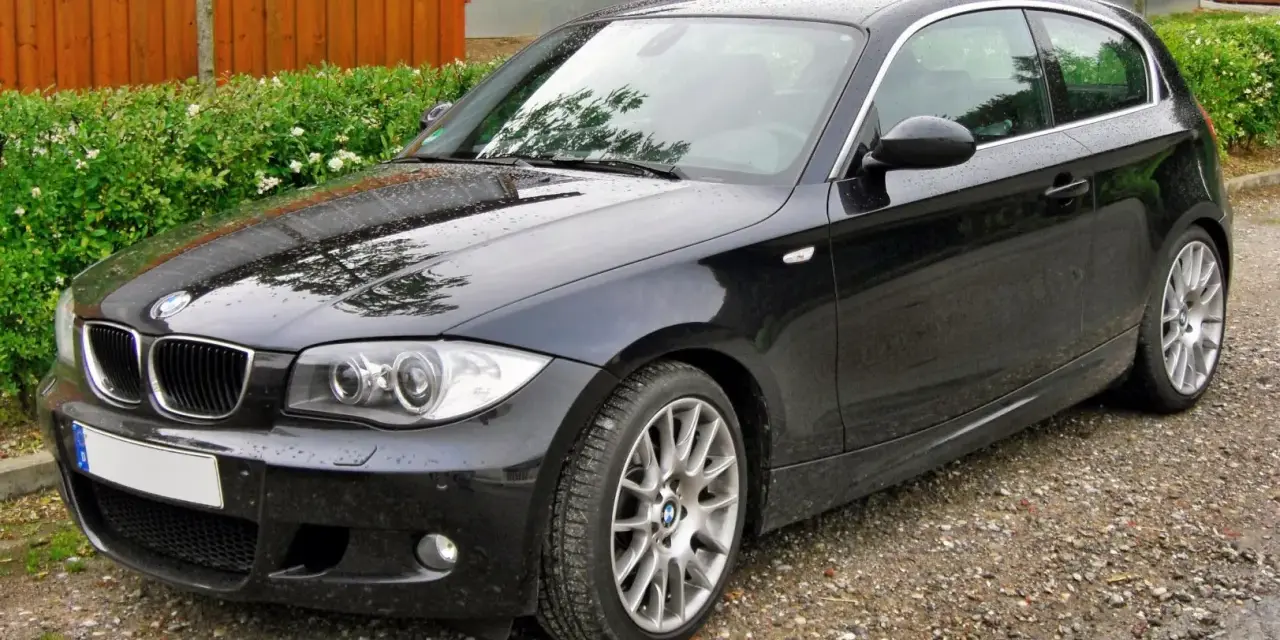 Czarny BMW e81, lśniący po deszczu, stoi na żwirowej drodze obok zielonego żywopłotu i drewnianego ogrodzenia.