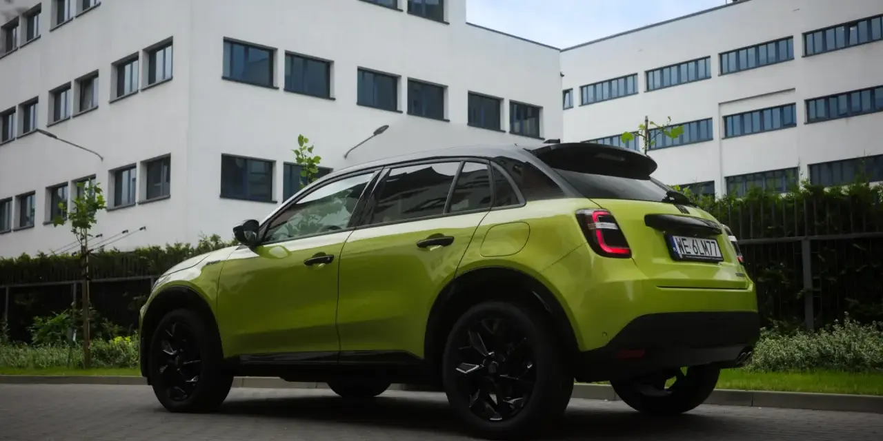 Jaskrawozielony Fiat 600 z czarnymi dodatkami parkuje przed nowoczesnym budynkiem.