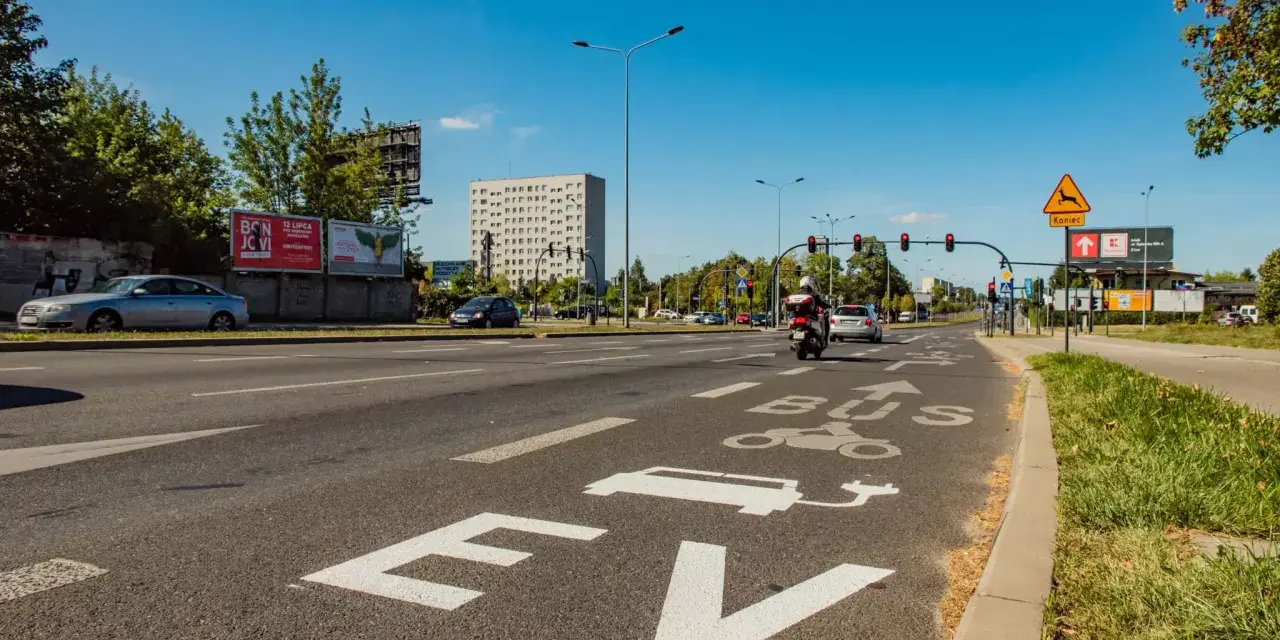 Na drodze oznaczenie "EV BUS" sugeruje, czy samochody elektryczne mogą jeździć buspasem. W tle budynek i ruch uliczny.
