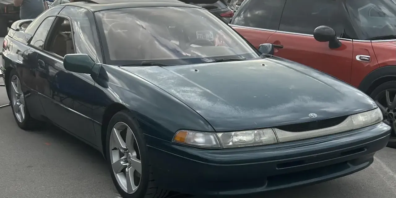Subaru SVX w ciemnozielonym kolorze, z charakterystycznymi oknami w stylu "glass-canopy", zaparkowany obok czerwonego samochodu.