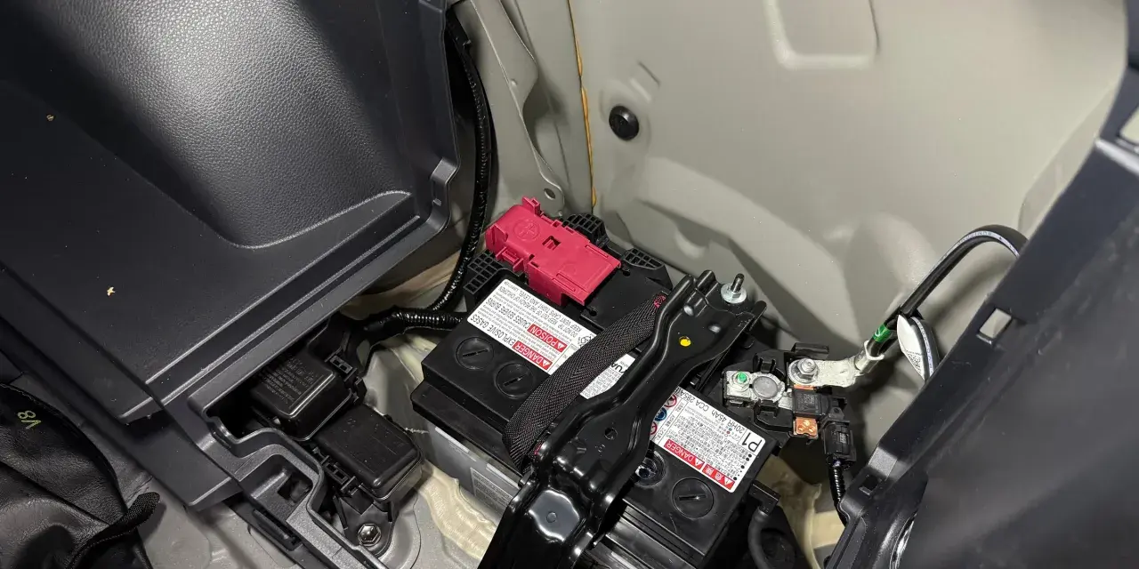 Akumulator 12V w Toyocie Prius znajduje się w bagażniku, pod plastikową osłoną. Widoczne są dwa akumulatory, jeden z czerwonym zabezpieczeniem.