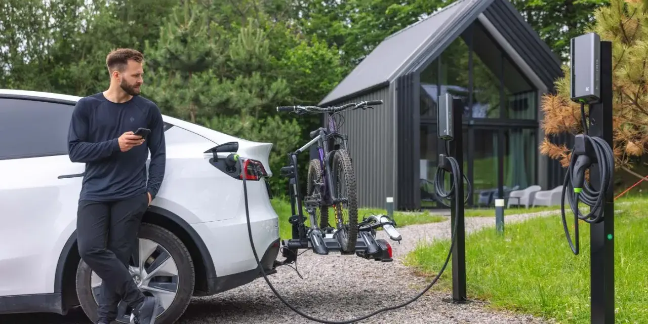 Mężczyzna z telefonem przy białym samochodzie elektrycznym, który ładuje się obok stojaka z rowerami. Obok stoją dwie ładowarki do samochodów elektrycznych.