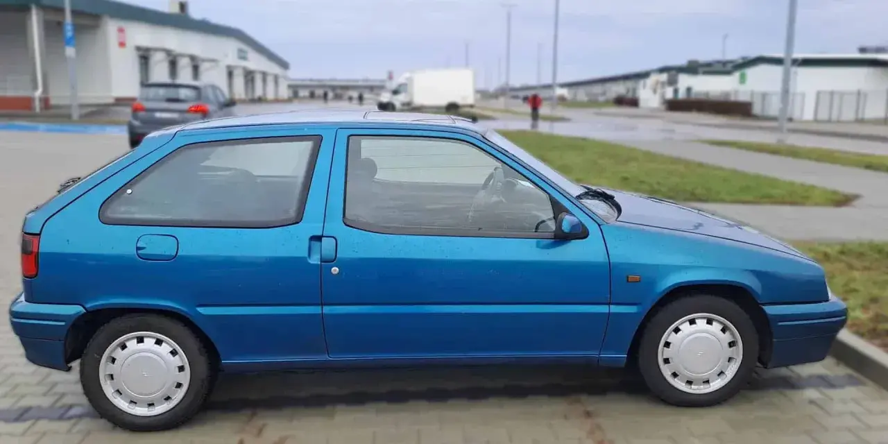 Niebieski Citroen ZX stoi na parkingu. W tle widać budynki i białą ciężarówkę.