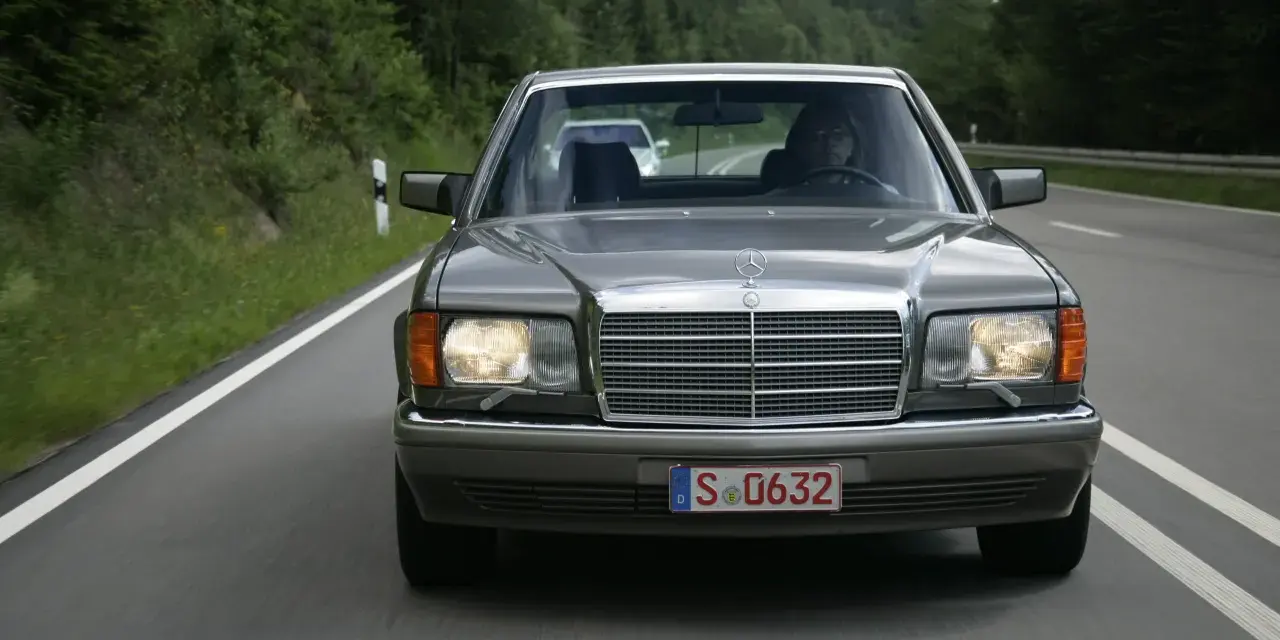 Szary Mercedes W126 jedzie drogą wśród zielonych lasów.