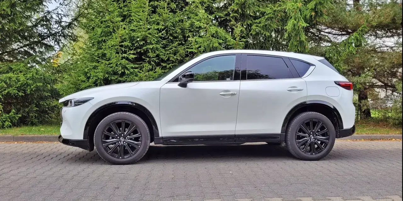 Nowa Mazda CX-5 w białym kolorze, zaparkowana na brukowanym podjeździe, otoczona zielenią drzew.
