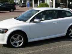 Białe Volvo C30 z felgami pięcioramiennymi stoi na parkingu przed salonem samochodowym.