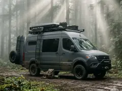 Szary bus z napędem 4x4, gotowy na przygodę w lesie. Na dachu bagażnik z wyposażeniem, z boku drabinka i koło zapasowe.