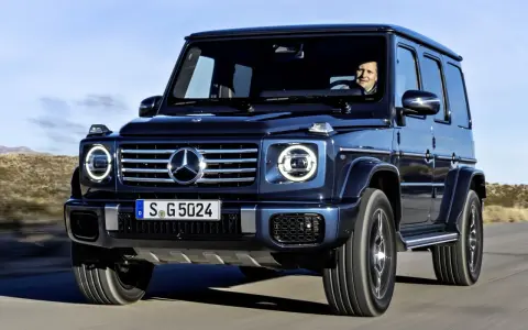 Ciemnoniebieski Mercedes G Klasa jedzie po pustynnej drodze. Kierowca uśmiecha się. Sprawdź mercedes g klasa cena!