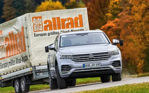 Srebrny Volkswagen Touareg holuje przyczepę z logo "Auto Bild allrad", jadąc po krętej drodze wśród jesiennych drzew.