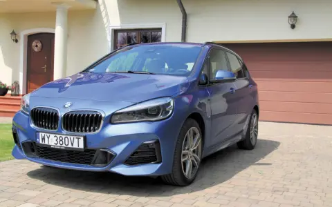 Niebieskie BMW 218i zaparkowane przed domem z brązową bramą garażową.