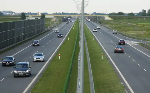 Na tej drodze szybkiego ruchu ruch jest umiarkowany. Widać kilka samochodów osobowych i jeden van. Po lewej stronie drogi znajduje się wysoki ekran akustyczny.