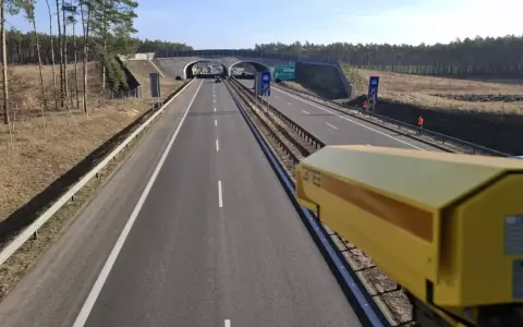 Żółta kamera monitoruje odcinek pomiarowy prędkości na S8. Droga prowadzi przez las, pod wiaduktem.