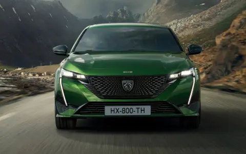 Zielony Peugeot 308 T7 jedzie drogą w malowniczym, górskim krajobrazie z widokiem na małą wioskę nad wodą.