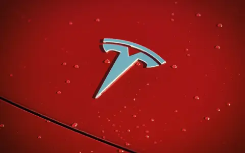 Błyszczące logo Tesli na czerwonej karoserii z kroplami deszczu. Czy Tesla jest tylko elektryczna?