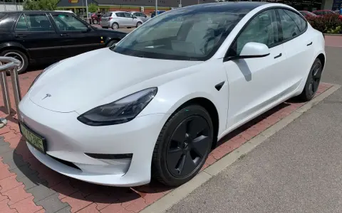 Biała Tesla Model 3 na parkingu przed sklepem. Zastanawiasz się, ile kosztuje najdroższa Tesla? Ten model to marzenie wielu kierowców.