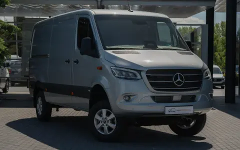 Srebrny bus Mercedes-Benz Sprinter, idealny jako jaki bus na budowę, stoi na parkingu przed nowoczesnym budynkiem.
