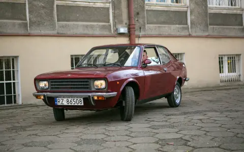 Czerwony fiat 128, klasyk z lat 70., zaparkowany na brukowanym dziedzińcu przed budynkiem.