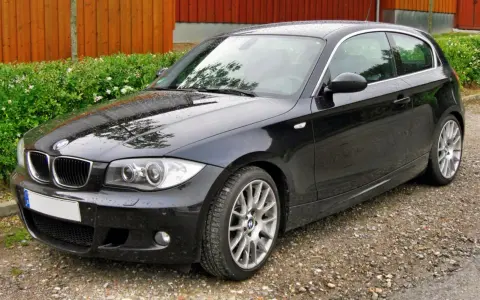 Czarny BMW e81, lśniący po deszczu, stoi na żwirowej drodze obok zielonego żywopłotu i drewnianego ogrodzenia.