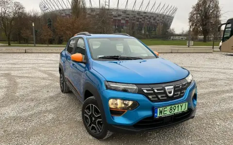 Niebieski Dacia Spring z pomarańczowymi akcentami na tle Stadionu Narodowego. Sprawdź atrakcyjną dacia spring cena!