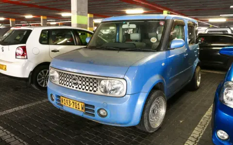 Niebieski Nissan Cube z charakterystycznym grillem i tablicą rejestracyjną N193-701 W, zaparkowany obok białego Volkswagena.