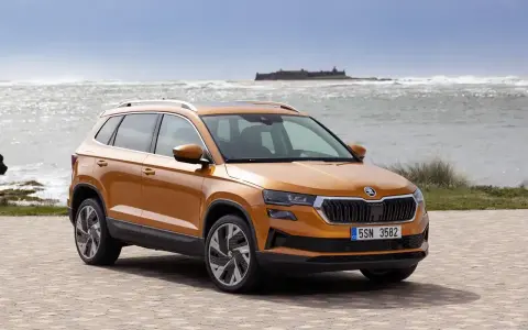 Pomarańczowa Skoda Karoq na tle morza i zamku. Sprawdź aktualny skoda cennik i wybierz swój wymarzony model.