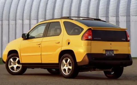 Żółty Pontiac Aztek z charakterystycznym tyłem i spojlerem, zaparkowany przed falistą, metalową ścianą.